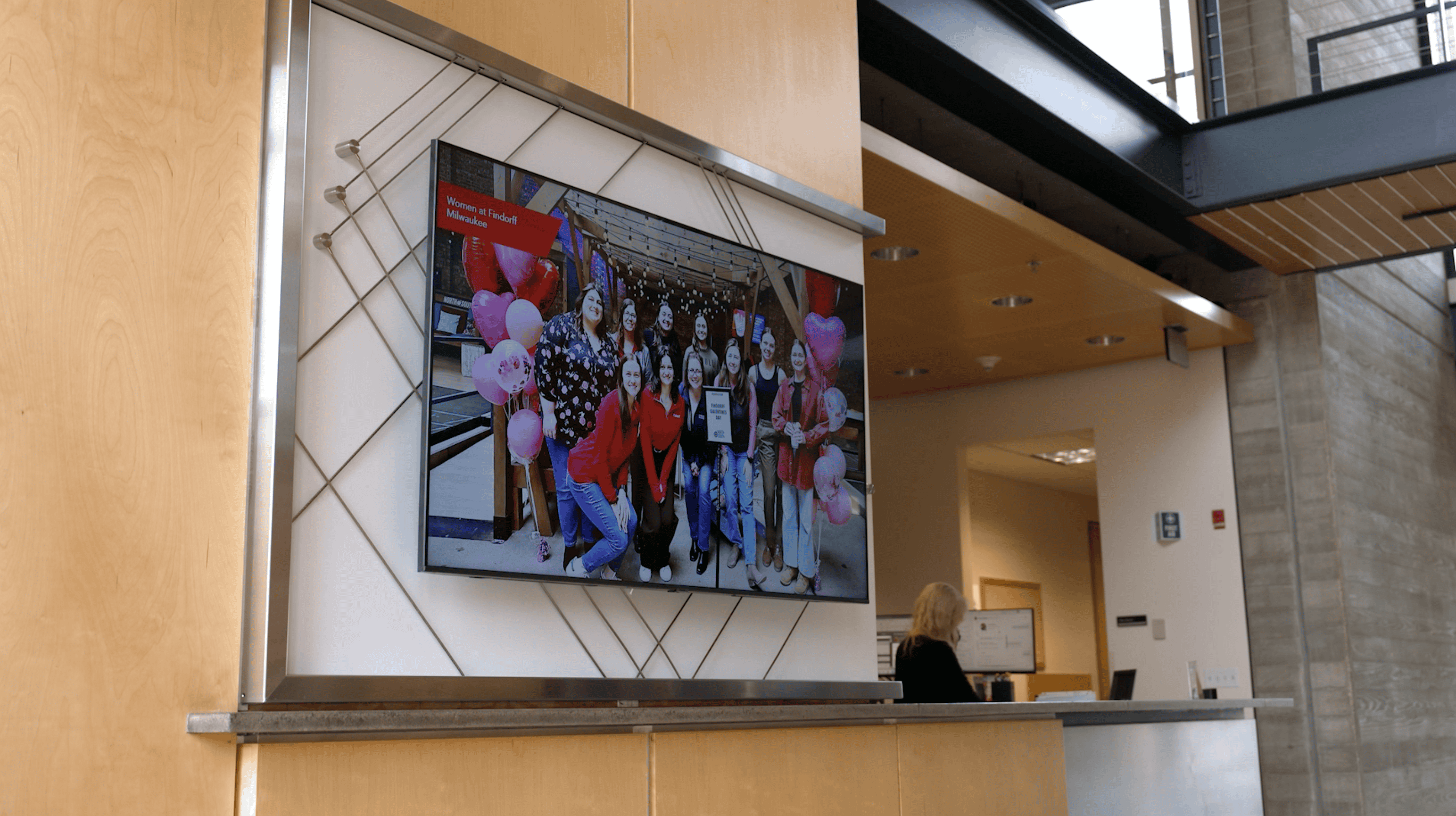 A digital display showing a group photo of people, with balloons in red and pink colors. The display is mounted within a metal-framed structure on a wooden wall, located near a reception desk in a modern office building.